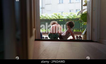 Kinder, die an der Tür sitzen und draußen Schuhe anziehen. Der ältere Bruder hilft dem jüngeren Geschwister, Schuhe an die Tür zu legen Stockfoto