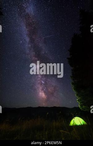 Sternenhimmel in den Bergen. Ein Zelt in der Nähe des Waldes wird von einer Taschenlampe beleuchtet. Milchstraße und Sternschnuppen am Nachthimmel. Camping-Leben. Stockfoto