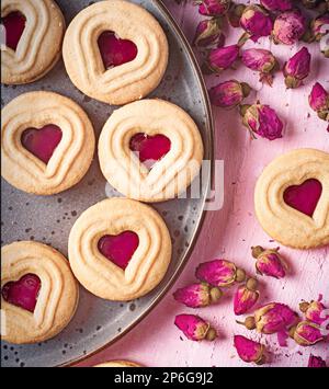 Vanillekekse mit herzförmigem Zentrum gefüllt mit Himbeeren auf rosa Hintergrund voller getrockneter Rosen Stockfoto