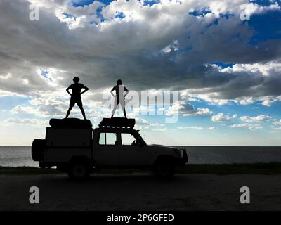 2 junge Erwachsene stehen auf einem Jeep als Silhouetten, die menschliche Figuren spielen. Sturmwolken am Himmel. Makgadikgadi Salzwannen, Botsuana, Afrika Stockfoto