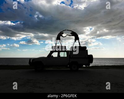 2 junge Erwachsene stehen auf einem Jeep als Silhouetten, die menschliche Figuren spielen. Sturmwolken am Himmel. Makgadikgadi Salzwannen, Botsuana, Afrika Stockfoto