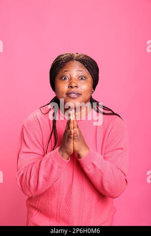 Eine afroamerikanische Frau, die Gebetshandsymbole im Studio trägt und religiöse spirituelle Geste auf pinkfarbenem Hintergrund zeigt. Ein junges Model mit Selbstvertrauen, das zu jesus für Frieden betet, christentum. Stockfoto