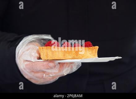 Nahaufnahme einer Hand mit einem Essenshandschuh, um ein Papierfach mit einer Obsttorte auf dem Prager Bauernmarkt am kalten Wintertag zu halten. Stockfoto