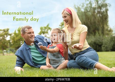 Glückliche Eltern und ihre Tochter blasen Seifenblasen im Park auf grünem Gras. Alles Gute Zum Familientag Stockfoto