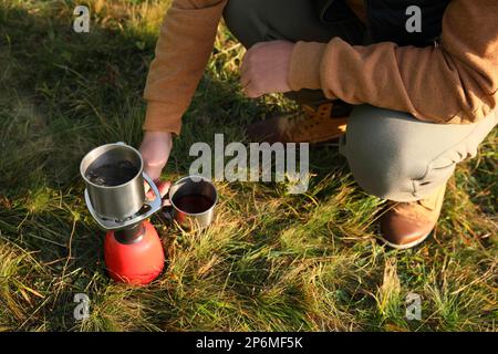 Ein Mann, der heißes Getränk macht, mit einem tragbaren Gasbrenner auf grünem Gras, Nahaufnahme. Campingsaison Stockfoto