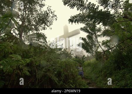 Ein hohes christliches Kreuz wird durch Nebel gesehen, im Vordergrund eines Dorfbewohners, der einen Pfad am Bukit Kasih (Hügel der Liebe) hinuntergeht, einem beliebten religiösen Reiseziel im Dorf Kanonang, West Kawangkoan, Minahasa, North Sulawesi, Indonesien. Gewidmet allen religiösen Gläubigen und Gläubigen, während sie die Geister der Liebe, des Friedens und der Toleranz fördern; Bukit Kasih wurde Anfang 2000 gegründet, als Adolf J. Sondakh Gouverneur der Provinz Nord-Sulawesi war. Es wurde als noble Friedensinitiative betrachtet. Stockfoto