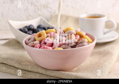 Gieße Milch in die Schüssel mit bunten Müsliringen auf Serviette vor weißem Grunge-Hintergrund Stockfoto