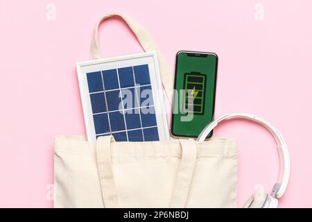 Tasche mit tragbarem Solarmodul, Mobiltelefon und Kopfhörern auf pinkfarbenem Hintergrund Stockfoto