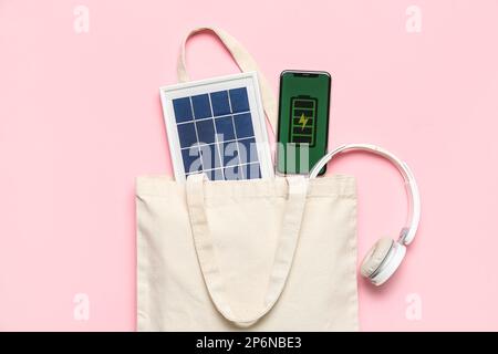 Tasche mit tragbarem Solarmodul, Mobiltelefon und Kopfhörern auf pinkfarbenem Hintergrund Stockfoto