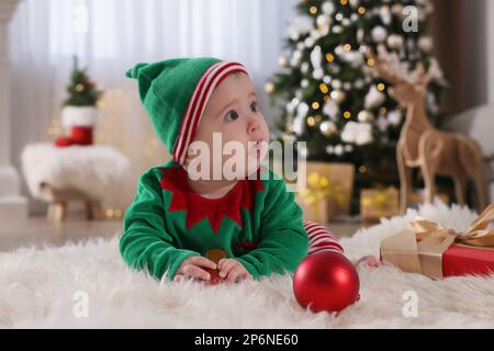 Baby trägt süßes Elfenkostüm auf dem Boden im Zimmer, das zu Weihnachten dekoriert ist Stockfoto