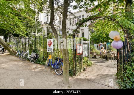 Fahrräder parken an einer Straßenseite vor einem Apartmentkomplex mit Bäumen und Menschen, die an Tischen sitzen Stockfoto