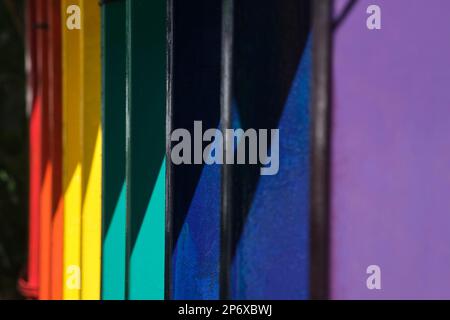 Regenbogenfarbener Zaun unter hartem Licht, parallele vertikale Linien und Diagonalen; helle und schattige Farbbereiche. Stockfoto