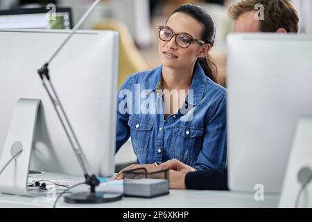 Sie zeigt ihrem Kollegen, wie es gemacht wird. Geschäftsfrau, die zwischen Computern gesehen wurde und ihrem Kollegen etwas zeigte. Stockfoto