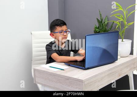 Ein 7-jähriger Latino-Junge mit Brille macht Hausunterricht und nimmt an Online-Kursen zu Hause mit einem Laptop Teil. Er lernt, ist überrascht und nimmt daran Teil Stockfoto