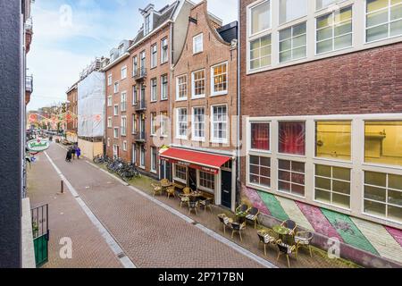 Amsterdam, Niederlande - 10. April 2021: Eine Straße mit Tischen und Stühlen in der Mitte, von einem überquerten Fenster aus gesehen Stockfoto
