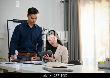 Zwei professionelle Geschäftsleute der Millennials in Asien schauen auf den Tablet-Bildschirm und diskutieren und diskutieren über ihr Finanzprojekt im Büro. Stockfoto