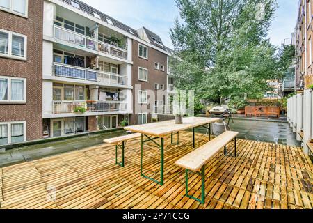 Amsterdam, Niederlande - 10. April 2021: Eine hölzerne Terrasse mit Bänken und Tischen vor einem Apartmentgebäude an einem sonnigen Tag, aus der Kamera aufgenommen Stockfoto