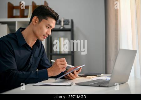 Professioneller und erfolgreicher asiatischer Geschäftsmann oder männlicher Manager, der an seinem Schreibtisch im Büro arbeitet und sein Unternehmen mit einem digitalen Tablet verwaltet Stockfoto