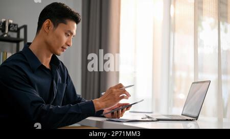 Seitliches Bild eines professionellen und attraktiven asiatischen Geschäftsmanns oder Geschäftsführers der Millennials, der sein Smartphone verwendet und SMS oder E-Mails sendet, während er vor ihm sitzt Stockfoto