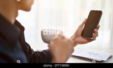 Zugeschnittenes und Nahaufnahme-Bild eines erfolgreichen und professionellen asiatischen Geschäftsmanns oder männlichen Chefs, der sein Telefon benutzt, während er an seinem Schreibtisch Kaffee trinkt i Stockfoto