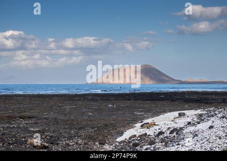 Dunkle Lava-Erde am Strand und weiße Korallenstücke. Ruhiger Atlantik. Vulkangebirge Lobos im Hintergrund. Fuerteventura, Kanarische Inseln, Stockfoto