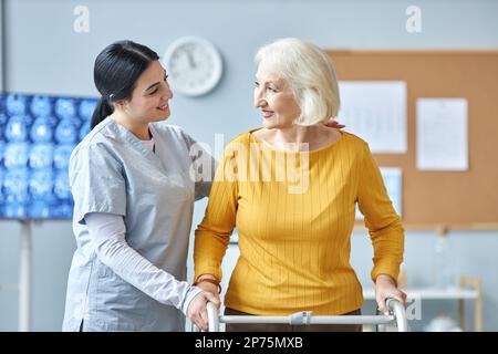 Taillenhochporträt einer lächelnden jungen Krankenschwester, die Seniorin mit Mobilitätswandler in der medizinischen Klinik hilft Stockfoto