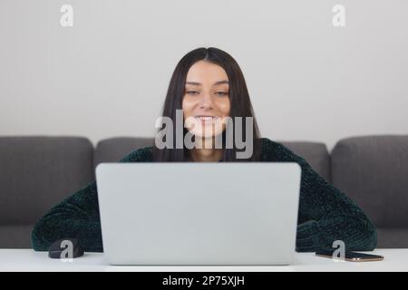 Fröhliche junge Frau, die in einer Videokonferenz spricht. Porträt einer schönen brünetten Frau, die mit einem Lächeln auf den Computerbildschirm schaut Stockfoto