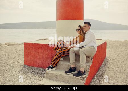 Ein Paar sitzt in der Nähe eines Leuchtturms am Strand vor dem Hintergrund des Meeres und der Berge. Urlaub, Flitterwochen, Reisen. Ein stilvolles Paar Stockfoto