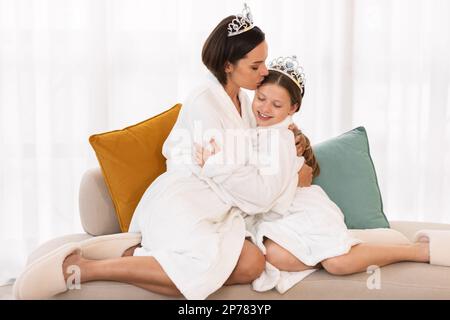 Mom Und Tochter Tragen Weiße Bademäntel Und Spielzeugkronen, Die Sich Zu Hause Umarmen Stockfoto