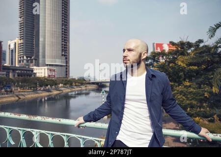Ernsthafter brutaler bärtiger Mann, der auf einer Brücke in einer Stadtlandschaft steht und denkt. Kahlköpfiger kaukasischer Kerl in lässiger Kleidung, deprimiert, wegblickend, alo Stockfoto