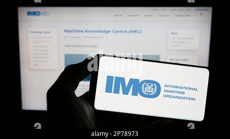 Person, die ein Mobiltelefon mit dem Logo der Internationalen Seeschifffahrtsorganisation (IMO) auf dem Bildschirm vor der Webseite hält. Konzentrieren Sie sich auf das Display des Telefons. Stockfoto