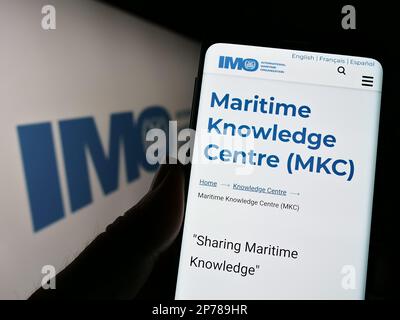 Person, die ein Mobiltelefon mit einer Webseite der Internationalen Seeschifffahrtsorganisation (IMO) auf dem Bildschirm vor dem Logo hält. Konzentrieren Sie sich auf die Mitte des Telefondisplays. Stockfoto