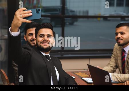 Freunde Geschäftspartner machen ein Selfie bei einem Meeting. Eine internationale Gruppe von Männern in Anzügen sitzt an einem Tisch im Freien und redet. Arbeitsmomente und Stockfoto