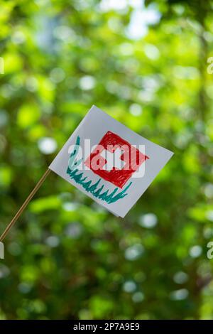 Kleinkind-Zeichnung der Schweizer Nationalflagge auf dem Papier. Nahaufnahme, geringe Schärfentiefe, grüner Naturhintergrund. Stockfoto