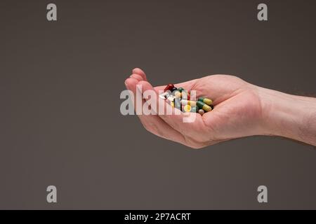 Weiße männliche Hand voll mit verschiedenen farbigen medizinischen Pillen, Tabletten und Kapseln. Nahaufnahme des Studios, isoliert auf braunem Hintergrund. Stockfoto