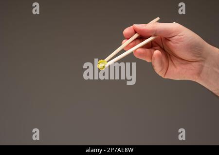 Frische grüne Wasabi-Paste auf Holzstäbchen, die von männlichen Weißen in der Hand gehalten werden. Nahaufnahme des Studios, isoliert auf Grau. Stockfoto