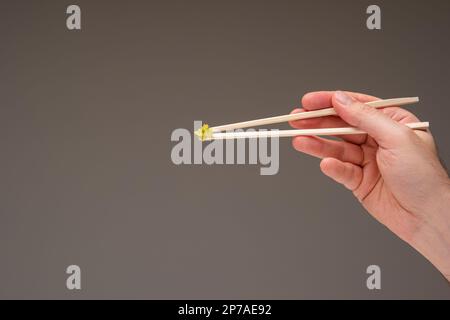 Frische grüne Wasabi-Paste auf Holzstäbchen, die von männlichen Weißen in der Hand gehalten werden. Nahaufnahme des Studios, isoliert auf Grau. Stockfoto