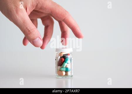 Kaukasische männliche Hand greift nach einer Glasflasche voll mit verschiedenen farbigen medizinischen Pillen, Tabletten und Kapseln. Nahaufnahme des Studios, isoliert auf Wai Stockfoto