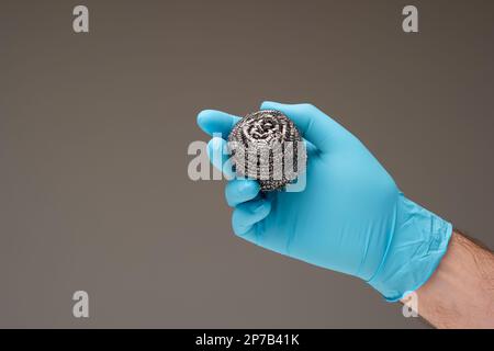 Ein neuer Küchenwäscher aus Metall, der von einem weißen Mann in der Hand gehalten wird und einen blauen Latexhandschuh trägt. Nahaufnahme des Studios, isoliert auf grauem Hintergrund. Stockfoto