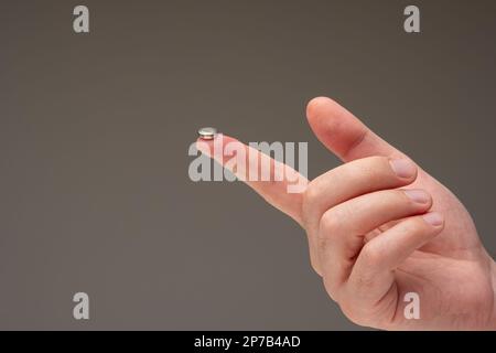 Sehr kleine L1131-Knopfzelle auf dem Zeigefinger eines kaukasischen Mannes. Nahaufnahme, isoliert auf grauem Hintergrund. Stockfoto