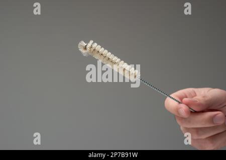Flaschenreinigungsbürste, die von der weißen männlichen Hand gehalten wird. Kunststoffbürste mit gewebtem Metallgriff, Nahaufnahme im Studio, isoliert auf grauem Hintergrund. Stockfoto