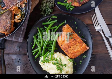 Gebratenes Lachsfilet in der Pfanne mit grünen Bohnen und Kartoffelpüree auf einem Teller auf einem Holztisch. Flach verlegt Stockfoto