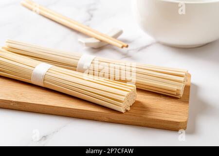 Getrocknete udon-Nudeln zum Kochen zubereitet. Rohe asiatische vegane Pasta aus Weizenmehl auf einem Bambusschneidbrett, Essstäbchen und einer Schüssel über der Marmorarbeitsfläche. Stockfoto