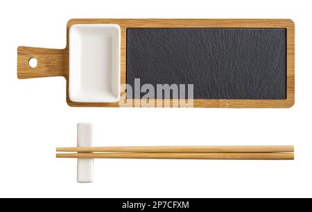 Bambus-Sushi-Servierset mit Ausschnitt. Holzbrett mit schwarzem Schieferblech, Sojasauce aus Porzellan und ein Paar Essstäbchen auf einem Rest isoliert auf einem weißen. Stockfoto