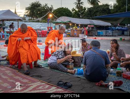 Eine Gruppe kambodschanischer Leute sitzt auf dem Boden, um auf dem Nachtmarkt in Phnom Penh, Kambodscha, zu essen, während zwei Buddhits Mönche zusehen. Stockfoto