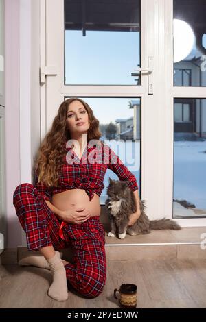 Eine schwangere Frau im Wohnzimmer mit einer Tasse Tee sitzt auf der Schwelle einer Glastür und spielt mit der Katze. Frau in rotem Karo-Pyjama Stockfoto