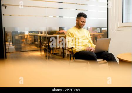 Konzentrierter, entschlossener, junger Freiberufler, der in einer entspannten Pose im Sessel sitzt, auf dem Laptop tippt, Programmierer arbeitet an einem Projekt, entwickelt Software, männliche Studenten websurfen Stockfoto