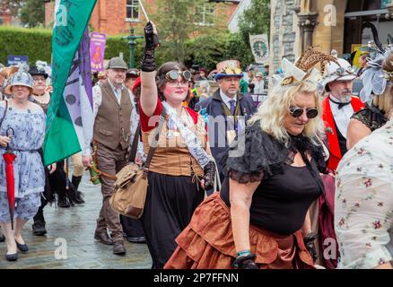 Eine Gruppe von Steampunken, die in einer Prozession entlang einer Straße laufen. Eine junge Frau, die eine Schärpe trägt, auf der steht "Stimmen für Frauen", die mit einer Flagge wedelt. Stockfoto