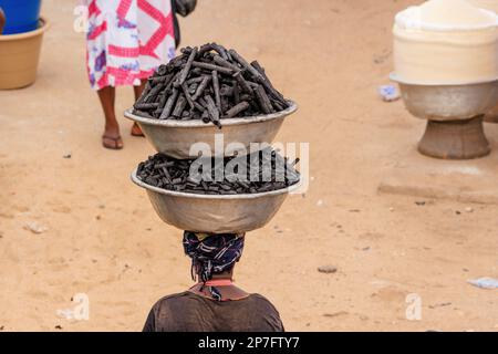 Rückansicht einer afrikanischen Dame, die zwei Metallschüsseln Holzkohle übereinander und dann auf ihrem Kopf balanciert Stockfoto