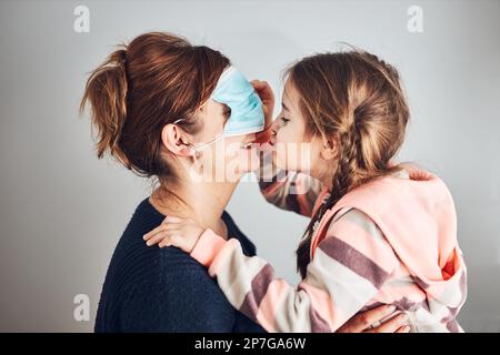 Kleines Mädchen, das die Gesichtsmaske ihrer Mutter auf die Augen bewegt und ihre Mutter küsst. Lustige Momente während der Quarantäne- und Covid-19-Pandemie Stockfoto